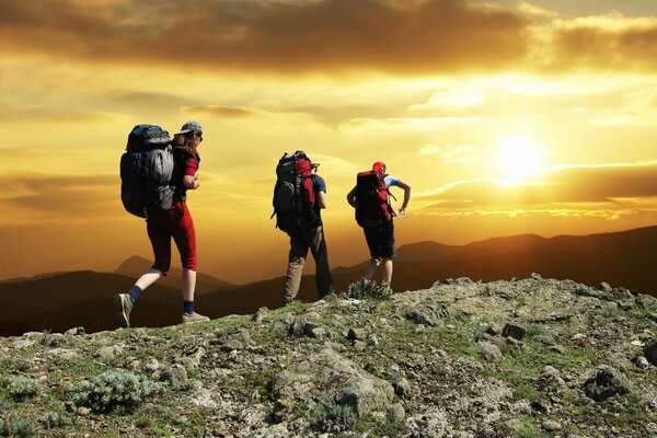 Three tourists with backpacks on the top of the mountain admire the setting sun