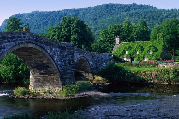 Eine Brücke über den Fluss und ein mit Grün bewachsenes Haus vor dem Hintergrund der Berge