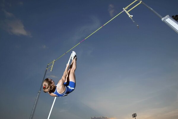 Atleta de uniforme azul que realiza un salto con pértiga durante una competencia al aire libre