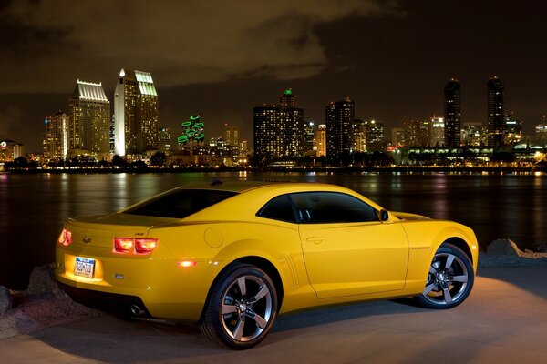 Żółty Chevrolet z włączonymi reflektorami na tle nocnego miasta