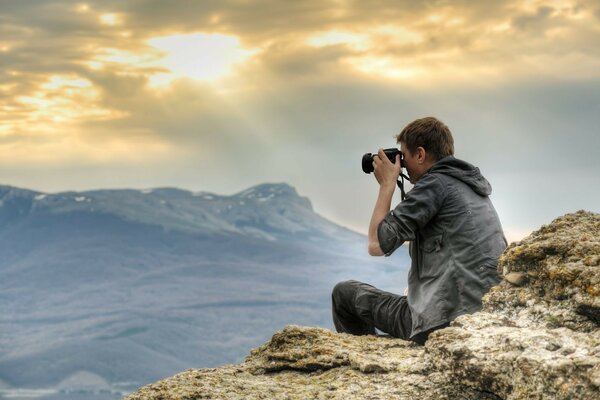 Der Kerl sitzt auf einem Felsen und filmt die Sonnenstrahlen, die durch die Wolken gehen, mit der Kamera