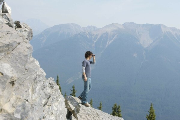 A man looks through binoculars from a cliff