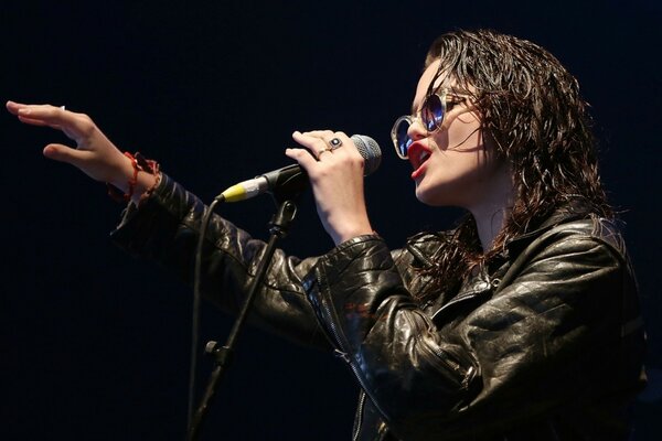 A dark-haired performer in a leather jacket and glasses sings into a microphone