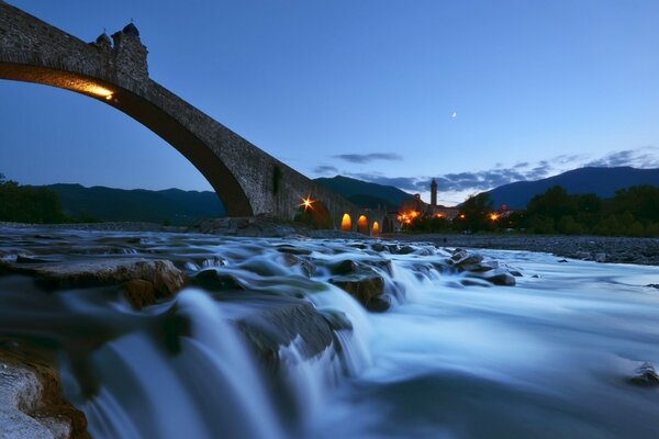 Boiling rocky river threshold in Italy with a bridge illuminated by yellow lights