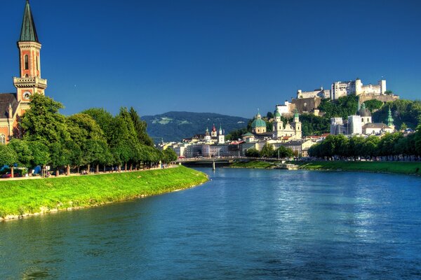 On both banks of the blue river there are churches and green trees against the blue sky