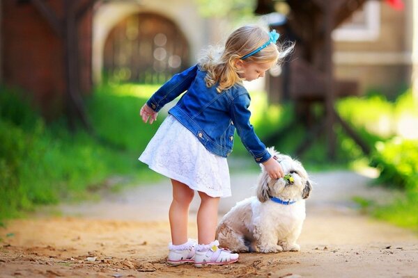 A little girl in a dress and a denim jacket strokes a fluffy dog