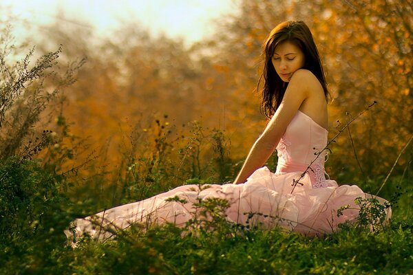 A brunette in a long pink dress is sitting in the grass against the background of an autumn forest