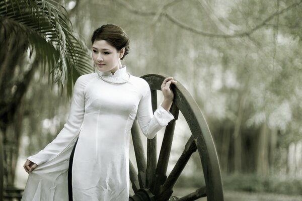 An Asian girl in a closed white dress rests her hand on a large wooden wheel