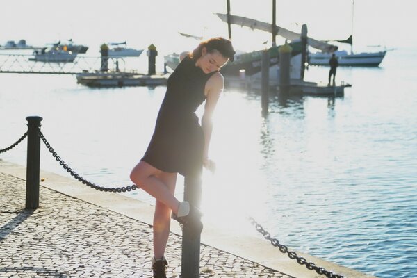 Una chica con un vestido corto en el muelle se apoya en un poste de metal