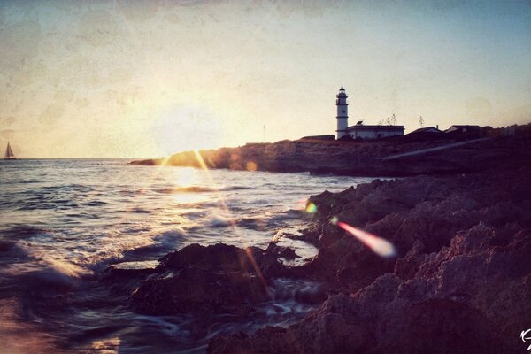 La costa del mar con un faro y un velero en el fondo del sol que se va