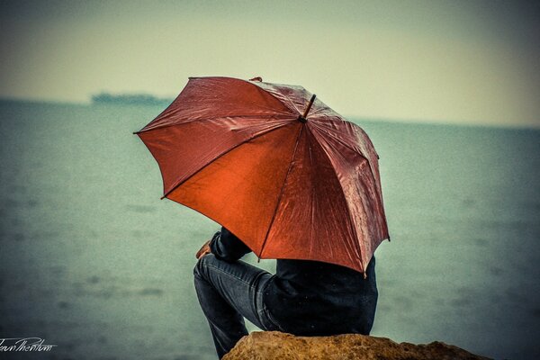 Homem sentado em uma pedra à beira-mar sob um guarda-chuva vermelho