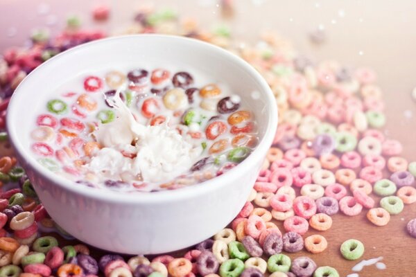 A white bowl with milk-soaked food on the table among the scattered multicolored cereals in the form of small circles