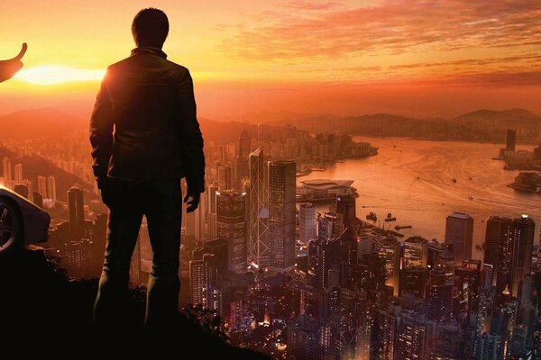 Against the background of the sunset sky, a man stands on a hill and looks at the city