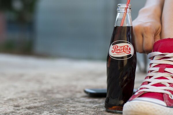 Someone is sitting relaxed in sneakers with a bottle of Pepsi Cola on concrete