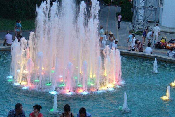 En una noche de verano, la gente se sienta en una fuente iluminada con diferentes colores