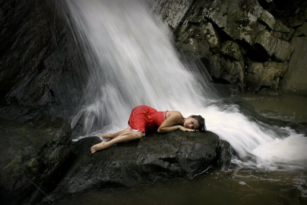 A girl is lying in a red dress on a stone next to a waterfall