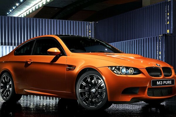 An orange BMW stands between shipping containers