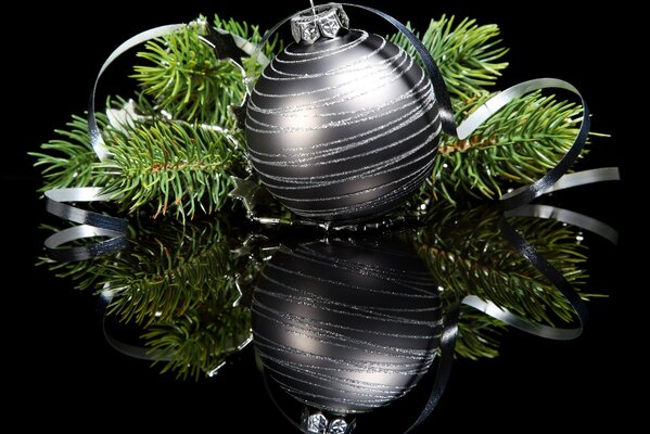 A silver ball with green Christmas tree branches on a black mirror