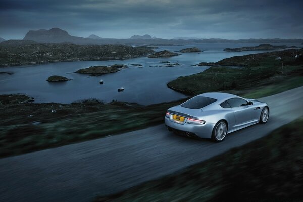 Une Aston Martin argentée longe le lac sur une route de campagne