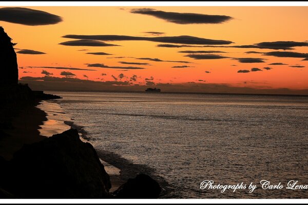 Red sunset, rocky coast, gray sea with a motor ship in the distance