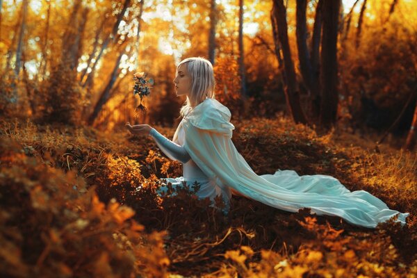 An elf holds a rose in his hands, sitting on the grass in the autumn forest