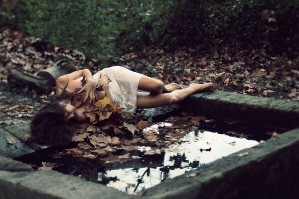 A girl in a nightgown is lying on the edge of the pool and covered with fallen leaves