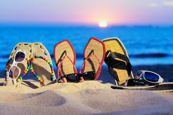 Chinelos, óculos na areia no fundo do pôr do sol do mar