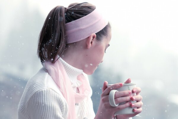 Una chica con una diadema rosa sostiene una taza en sus manos contra un fondo de nieve fina