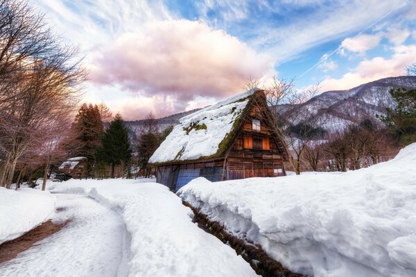 Dağın eteklerinde çatıyla karla kaplı küçük bir ev ve beyaz bulutlarla mavi bir gökyüzü